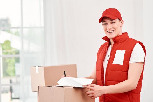 Portrait of delivery man in red postal uniform standing near stack of boxes, studying the list of clients for today. Friendly worker, high quality delivery service. Indoors.