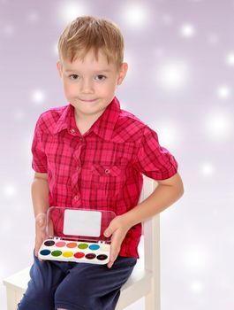 The concept of celebrating the New year, Holy Christmas, or child's birthday on a purple background and white snowflakes.Cute little boy in a red shirt holds his box of watercolors.