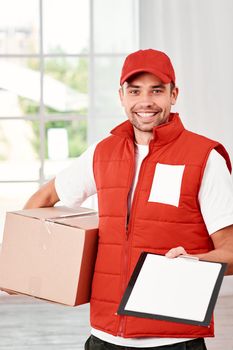 Cheerful postman wearing red postal uniform is delivering parcel to a client. He holds carton box, waiting for a signature on delivery and looking at the camera with a smile. Friendly worker, high quality delivery service. Indoors. Bright interior.