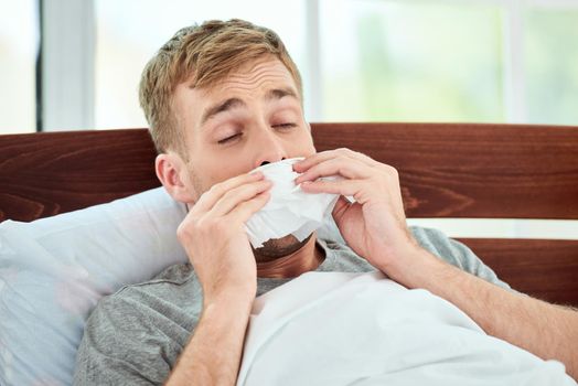 Portrait of a sick young man with runny nose suffering from cold or flu and sneezing while lying in bed at home. Virus disease. Coronavirus concept. Healthcare concept