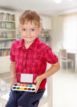The concept of family happiness,and preschool education of the child , against a child's room with bookshelves.Cute little boy in a red shirt holds his box of watercolors.
