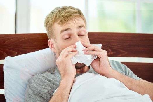 Portrait of a sick young man with runny nose suffering from cold or flu and sneezing while lying in bed at home. Virus disease. Coronavirus concept. Healthcare concept