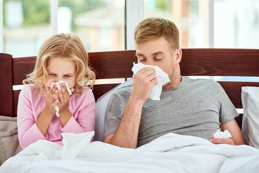 Father and daughter suffering from flu or cold and wiping noses while lying in bed together at home. Virus disease. Coronavirus concept. Sick family at home. Health concept