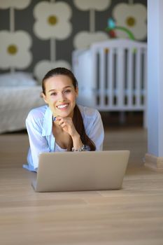 Young woman sitting on the floor near children's cot with laptop. Young mom.