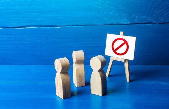 A group of people figurines looking at an easel with a red prohibitory symbol NO. Expression of protest and disagreement. Inaccessibility and absence. Quarantine and lockdown. Hard restrictions