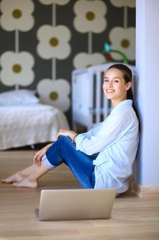 Young woman sitting on the floor near children's cot with laptop. Young mom.