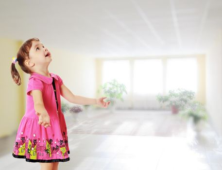 Adorable little girl with pigtails on the head , in a pink dress. The girl was looking at the top turned sideways to the camera.Against the background of a large room with Windows and flowers .