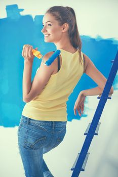 Happy beautiful young woman doing wall painting, standing near ladder.