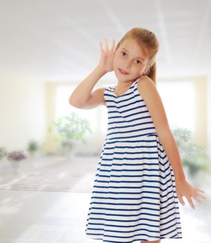 Caucasian little girl in a striped summer dress, listening. Girl holding a hand to his ear.On the background of the school hall with large Windows.