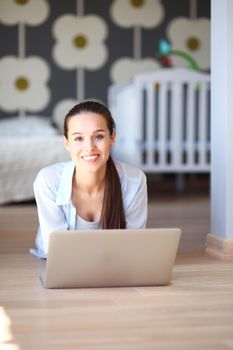 Young woman sitting on the floor near children's cot with laptop. Young mom.