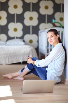 Young woman sitting on the floor near children's cot with laptop. Young mom.