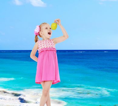 Little girl in a pink dress bites off an apple on a background of the sea.