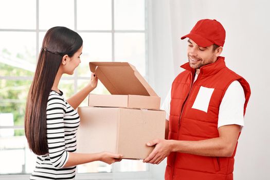 Cheerful postman wearing red postal uniform is delivering parcels to a satisfied client. He brings carton boxes to its owner. Dark-haired woman opens parcel, she is glad to receive it. Friendly worker, high quality delivery service. Indoors. Bright interior.