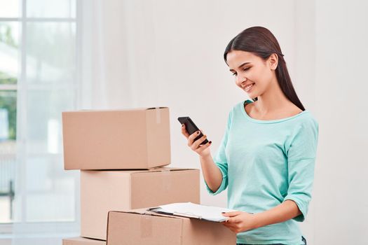 Young entrepreneur, SME, freelance woman working online, business by using smartphone, making purchase, order and preparing packages. Woman standing near stack of boxes, holding a clipboard and looking at her phone