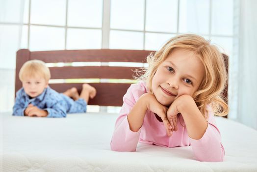 Cute little sister and her brother in pajamas lying on a big white mattress and looking at camera with smile. Quarantine. Bedroom. Family