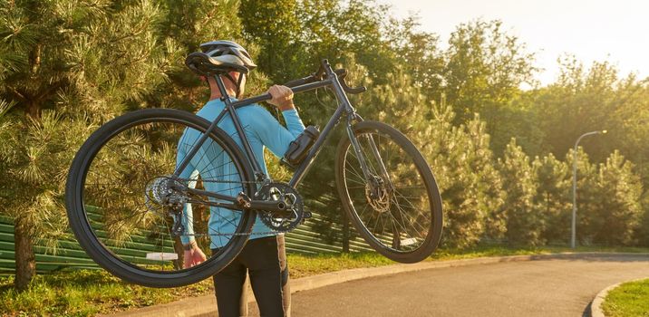 Strong athletic man in sportswear holding a bicycle while standing in park at sunset, cycling outdoors and enjoying amazing nature view. Active lifestyle and sport