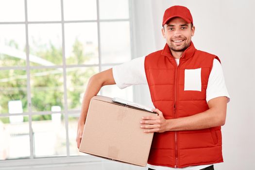 Cheerful postman wearing red postal uniform is delivering parcel to a client. He holds carton box and looking at the camera with a smile. Friendly worker, high quality delivery service. Indoors. Bright interior.