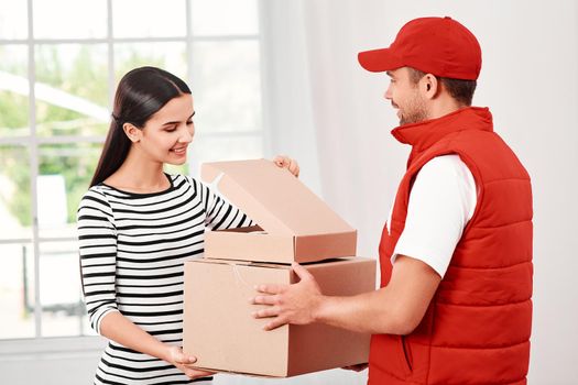 Cheerful postman wearing red postal uniform is delivering parcels to a satisfied client. He brings carton boxes to its owner. Dark-haired woman opens parcel, she is glad to receive it. Friendly worker, high quality delivery service. Indoors. Bright interior.