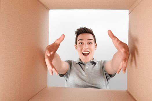 Cheerful boy wearing grey T-shirt unpacking, opening carton box. He stretches his arms forward to get a purchase out of the box. He is delighted with the speed of delivery of goods