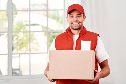 Cheerful postman wearing red postal uniform is delivering parcel to a client. He holds carton box and looking at the camera with a smile. Friendly worker, high quality delivery service. Indoors. Bright interior.