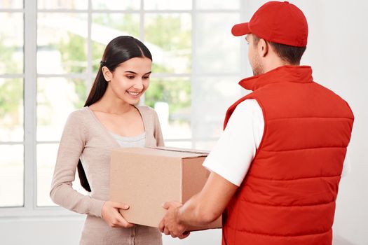 Cheerful postman wearing red postal uniform is delivering parcel to a satisfied client. He brings carton box to its owner. Dark-haired woman is taking parcel, she is glad to receive it. Friendly worker, high quality delivery service. Indoors. Bright interior.