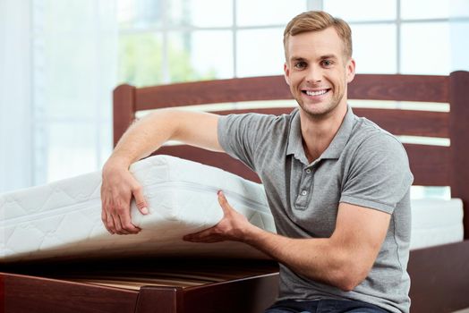Young and happy handsome man showing big and comfortable ergonomic mattress at camera and smiling. Bedroom. Home. Healthcare