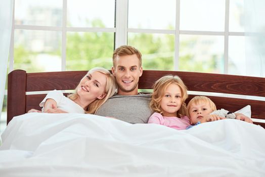 Young beautiful family of four people relaxing in bed together and looking at camera with smile. Quarantine. Enjoying weekend at home. Parents and kids