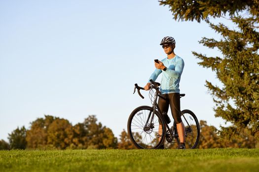 Taking a break. Young athletic man, professional cyclist in sportswear and protective helmet standing with his bike in park, using his smartphone and checking fitness results. Active lifestyle