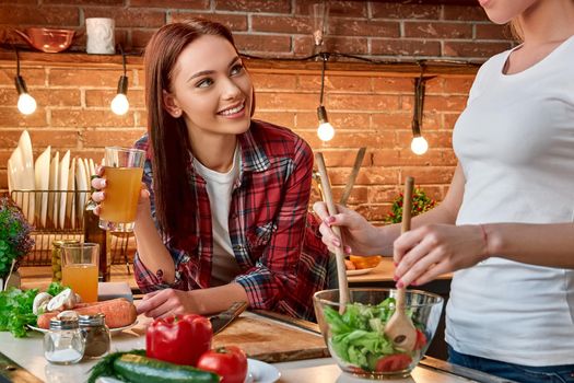 Portrait of two women, having fun, while preparing salad. They are fully involved in the process. Girl in white T-shirt mixes chopped vegetables in a bowl and her dark-haired friend in checkered shirt supervises the cooking process, drinking fresh juice