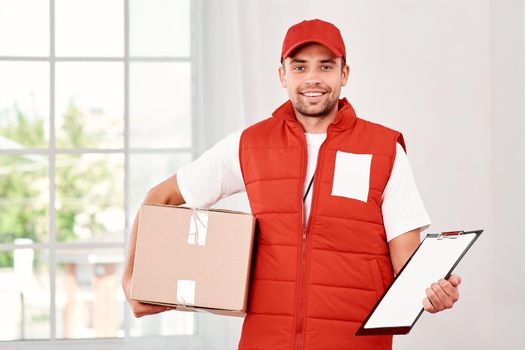 Cheerful postman wearing red postal uniform is delivering parcel to a client. He holds carton box, folder and looking at the camera with a smile. Friendly worker, high quality delivery service. Indoors. Bright interior.