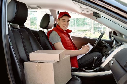 Parcel delivery man in red postal uniform drives a car. He is looking at the camera with a smile. Transport postal delivery courier delivering packages to customers. Impressive service.