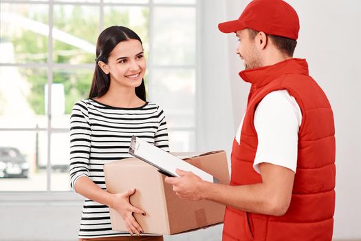 Cheerful postman wearing red postal uniform is delivering parcel to a satisfied client. He brings carton box to its owner. Dark-haired woman is taking parcel, she is glad to receive it. Friendly worker, high quality delivery service. Indoors. Bright interior.