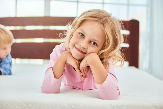 Portrait of a cute little girl in pink pajama smiling at camera while lying with her brother on a big and soft white comfortable mattress. Quarantine. Bedroom. Family