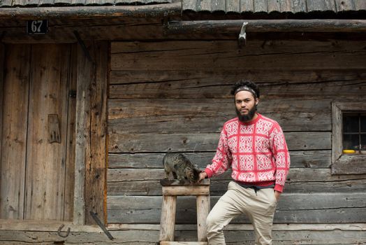 portrait of young hipster man with beard and cat in front of wooden house