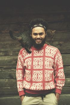 portrait of young hipster man with beard and cat in front of wooden house