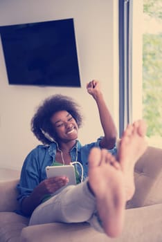 young beautiful African American woman enjoys listening to music with headphones and tablet in your armchair in her luxury home