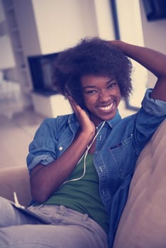 young beautiful African American woman enjoys listening to music with headphones and tablet in your armchair in her luxury home