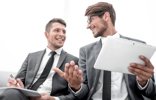 two young people are sitting in the office. One holds a tablet and gesticulates. The other holds a paper and a pen and listens.