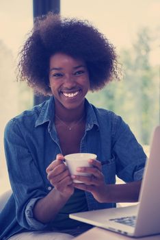 Young african american woman smiling sitting near bright window while looking at open laptop computer on table and holding white mug in her luxury home