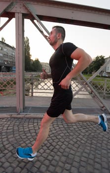 young sporty man jogging across the bridge at sunny morning in the city