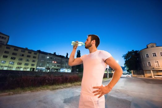 young athletic man drinking water after a night running session in the city