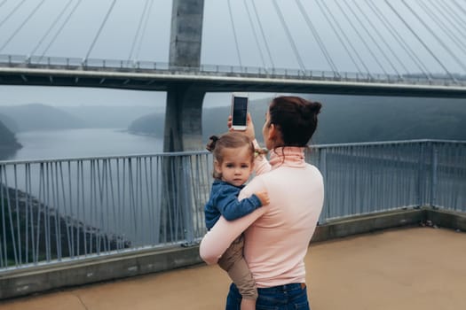 Mother and little daughter traveling together Mother photographing bridge while little girl sitting on hands France Bresta city. Caucasian family with one child standing observation deck take a photo