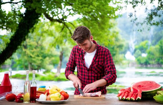 young man cutting raw meat for barbecue grill during outdoor french dinner party near the river on beautiful summer evening in nature
