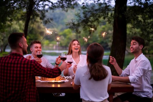 group of happy friends celebrating holiday vacation using sprinklers and drinking red wine while having picnic french dinner party outdoor near the river on beautiful summer evening in nature