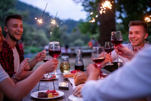 group of happy friends celebrating holiday vacation using sprinklers and drinking red wine while having picnic french dinner party outdoor near the river on beautiful summer evening in nature