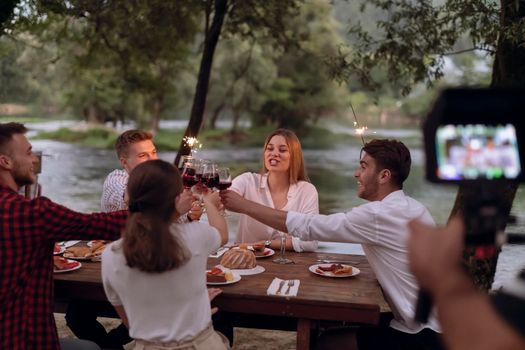 group of happy friends toasting red wine glass while having picnic french dinner party outdoor during summer holiday vacation near the river at beautiful naturethe cameraman records a group of friends having lunch and having fun on the river bank