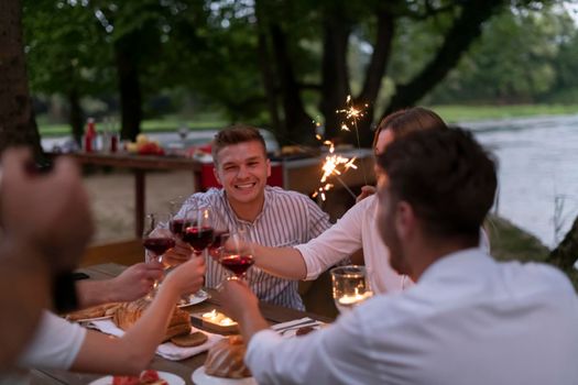 group of happy friends toasting red wine glass while having picnic french dinner party outdoor during summer holiday vacation near the river at beautiful naturethe cameraman records a group of friends having lunch and having fun on the river bank