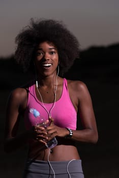 portrait of a young beautiful african american woman with headphones and a bottle of water after jogging in nature beautiful summer night