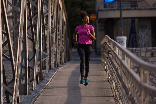 Young sporty african american woman running on sidewalk across the bridge at early morning jogging with city sunrise scene in background