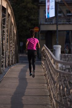 Young sporty african american woman running on sidewalk across the bridge at early morning jogging with city sunrise scene in background